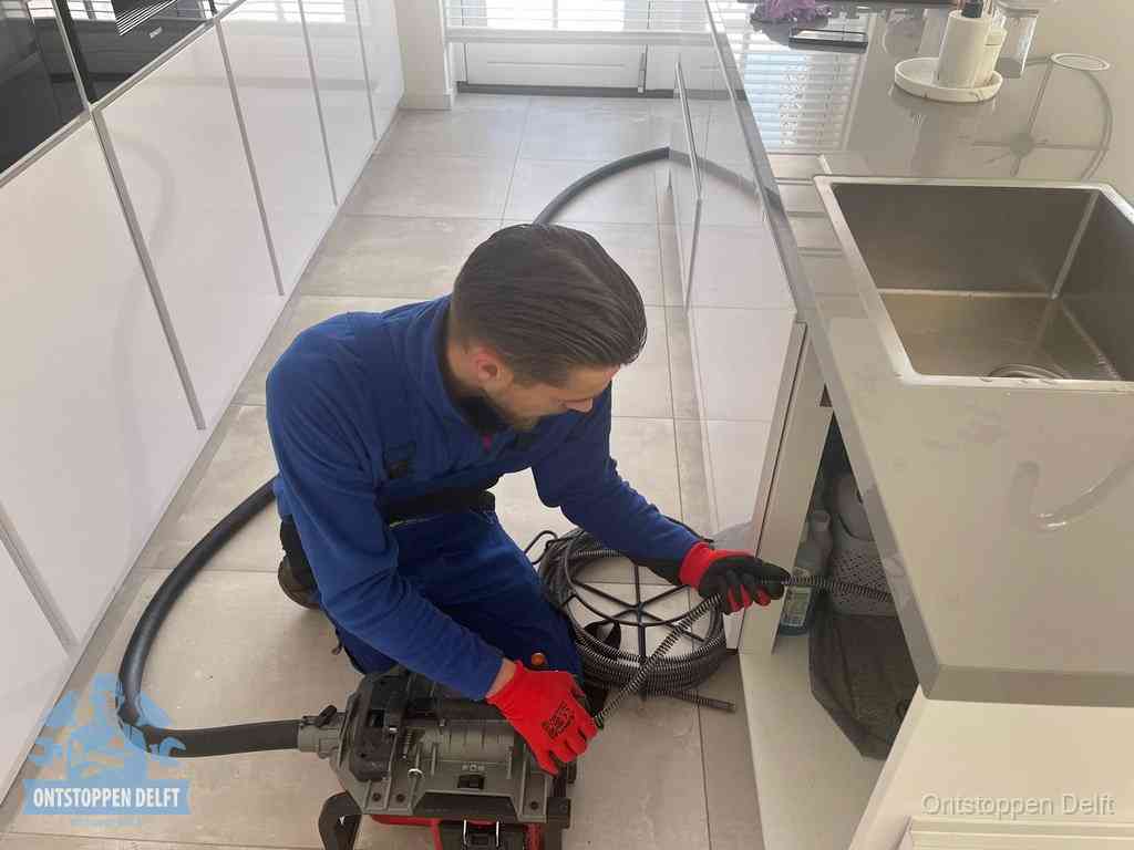 Een monteur ontstopt de keukenafvoer onder de spoelbak met een elektrische veermachine in Delft