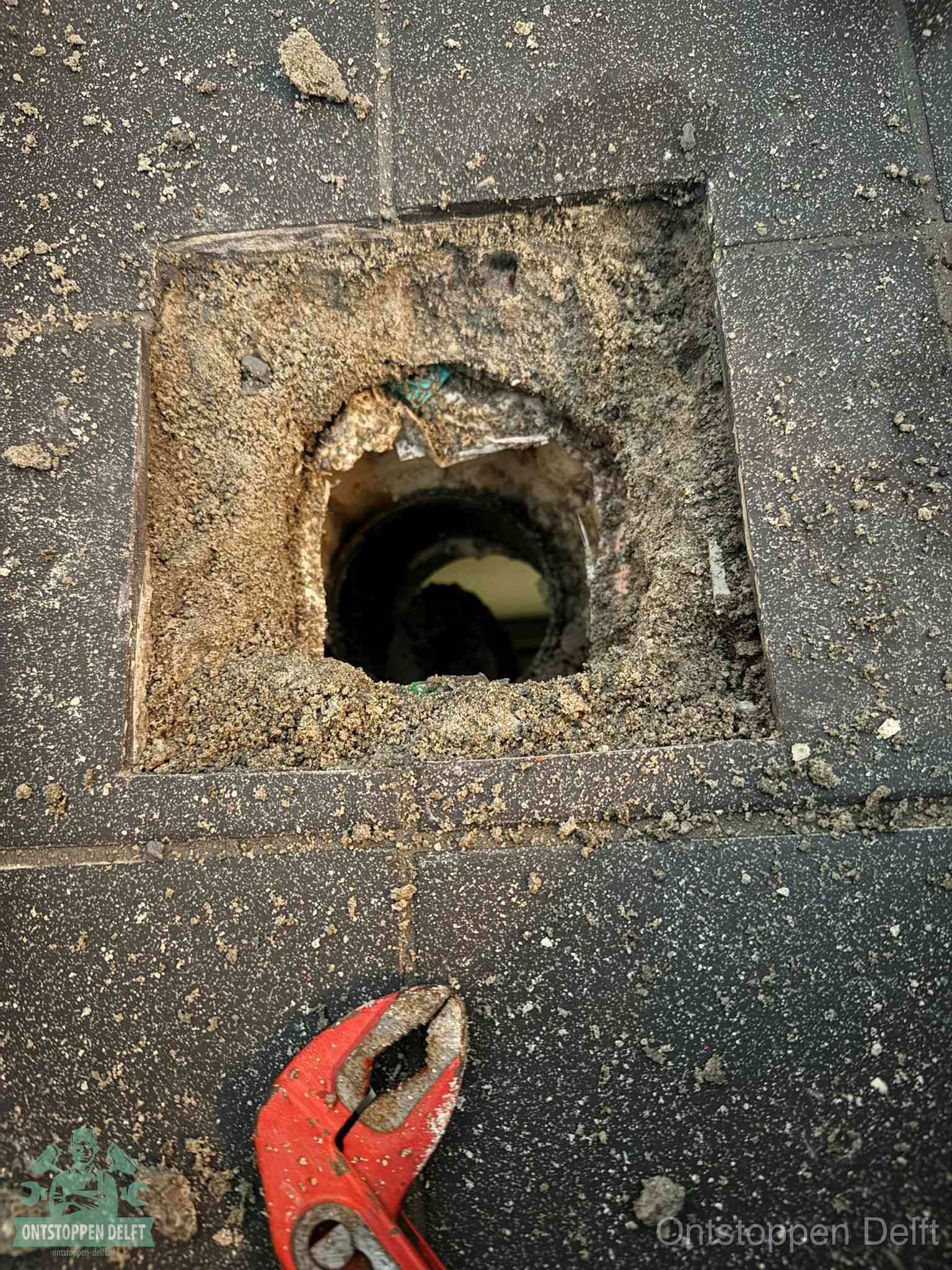 Een geopend rioolputje in een betontegelvloer met losgemaakt rooster en een zichtbare ronde afvoerbuis, het achterhalen waar de afvoerverstopping zit in Delft