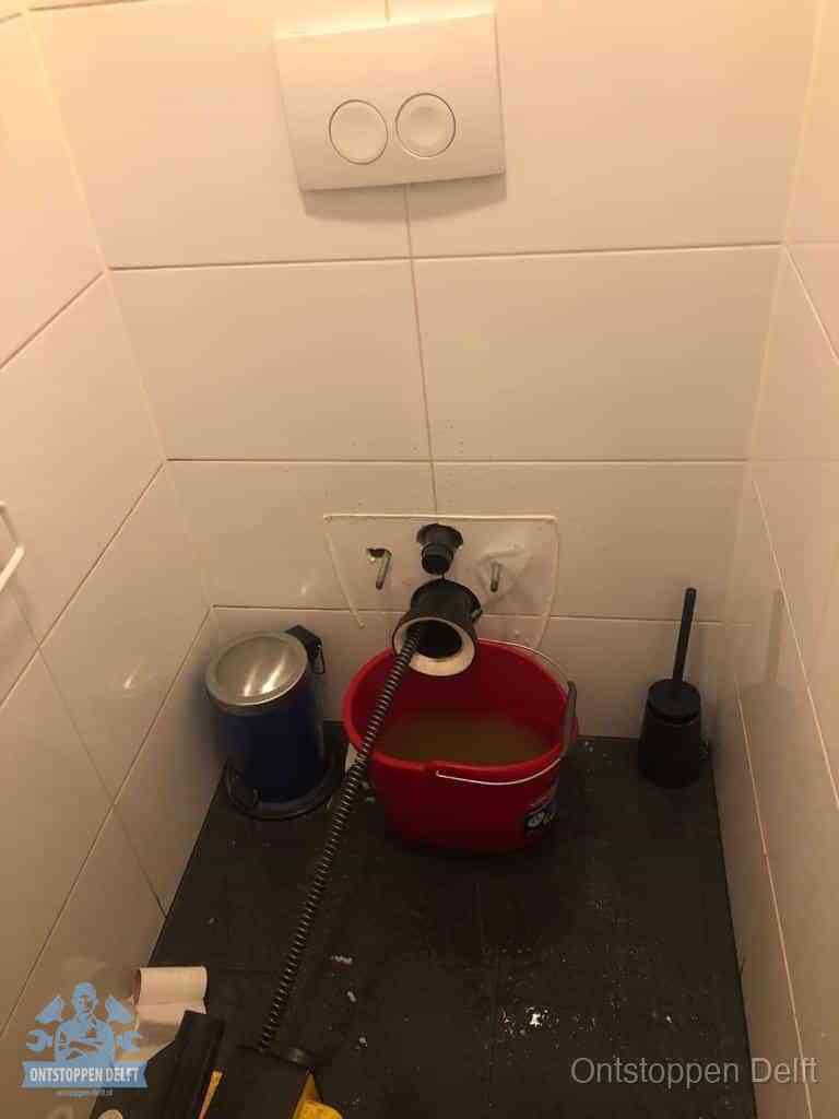Een ontstopte wand-wc met verwijderde toiletpot waar een ontstoppingsveer in de wanduitgang verdwijnt, het achterhalen waar de verstopping zit in Delft