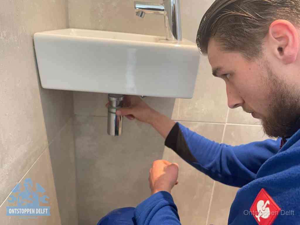 Een monteur draait met de hand de zwanenhalssifon onder een fonteinwasbak in de toiletruimte los om de bron van rioollucht in Delft te vinden