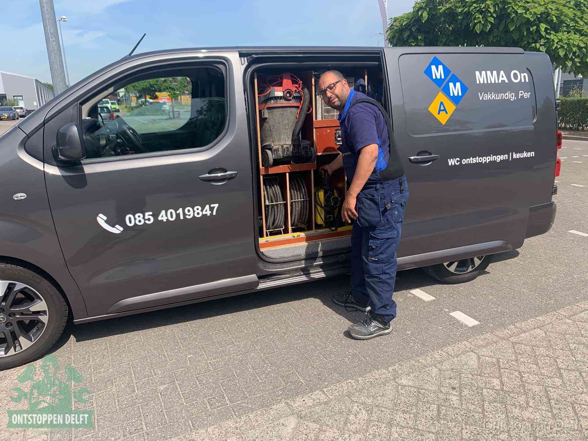 Een monteur bij de open schuifdeur van zijn bedrijfsbus op een parkeerplaats, klaar om de oorzaak van terugkerende afvoerverstoppingen aan te pakken in Delft