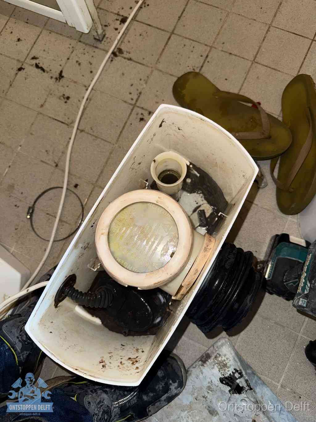 Een gedemonteerde toiletpot op de vloer met ontstoppingsveer en gereedschap ernaast, het verwijderen van een vastzittend voorwerp uit de wc in Delft