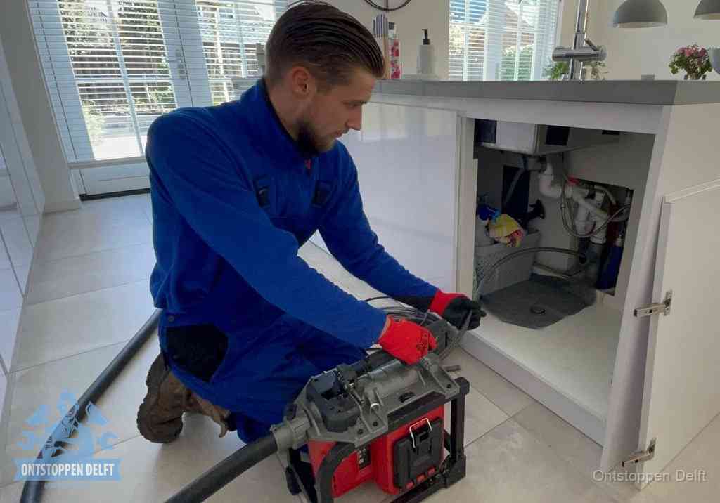 Een monteur voert een professionele elektrische ontstoppingsmachine in de afvoer onder de spoelbak tijdens het gootsteen ontstoppen in Delft